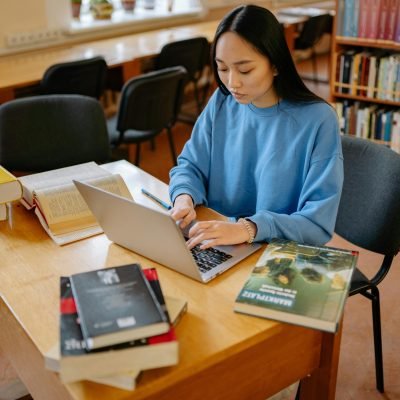 pexels-photo-8199252-8199252 Asian woman typing on a laptop surrounded by books in a library setting.